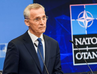 Man in a dark suit and blue tie speaks at a podium, with a NATO emblem and flag visible in the background.