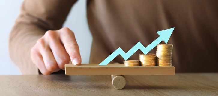 A person balancing stacked coins on a wooden plank with an upward arrow, symbolising CFD leverage and the potential for amplified gains or losses in trading.