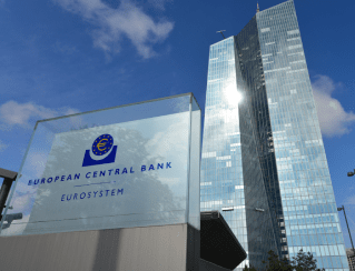 European Central Bank sign in front of tall glass office buildings on a sunny day, blue sky above.