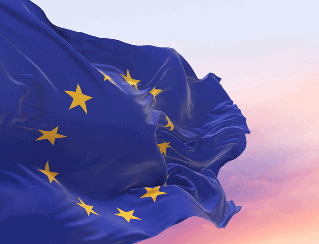 European Union flag with 12 gold stars on a blue field fluttering in a pink sunset sky.