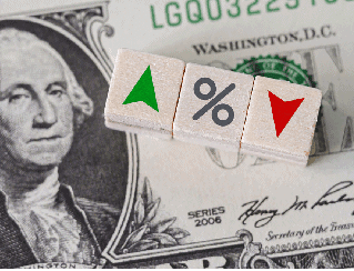 Close-up of a dollar bill with three wooden blocks showing an up green triangle, a percent sign, and a down red triangle, suggesting changing rates.
