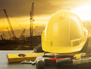 Yellow hard hat and scattered tools on a wooden surface with a construction site and cranes silhouetted at sunset in the background.