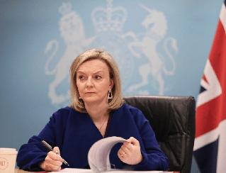 Female government official seated at a desk, signing a document with a British flag and royal crest backdrop behind her.