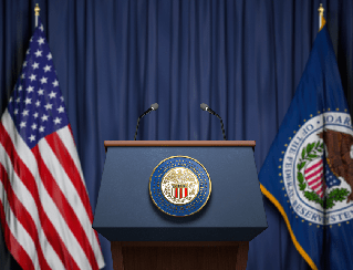 Podium with the U.S. presidential seal, flanked by American and government flags on a blue-curtained stage.