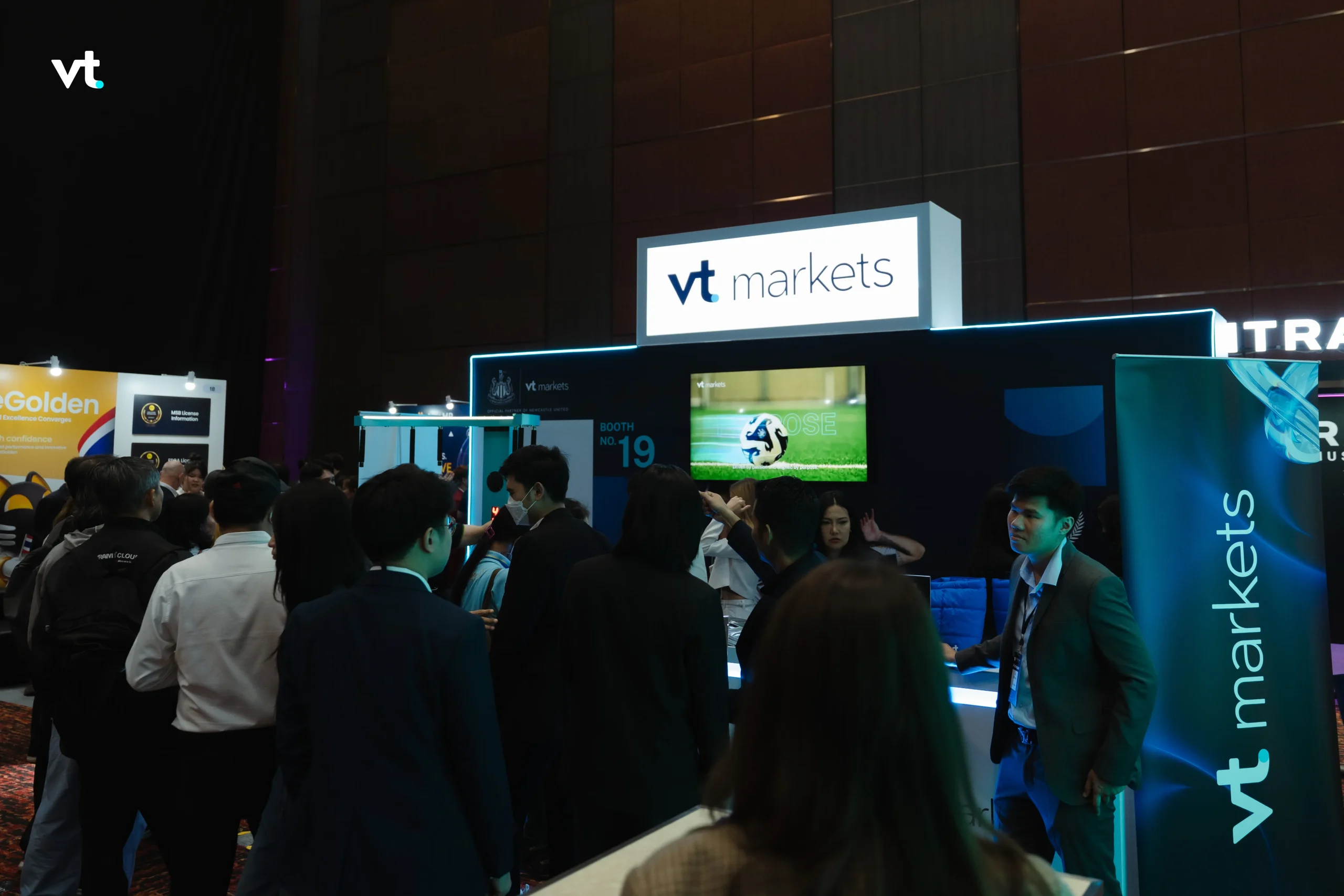 Busy convention floor with people gathered around a VT Markets booth and glowing signage on dark walls in the background.