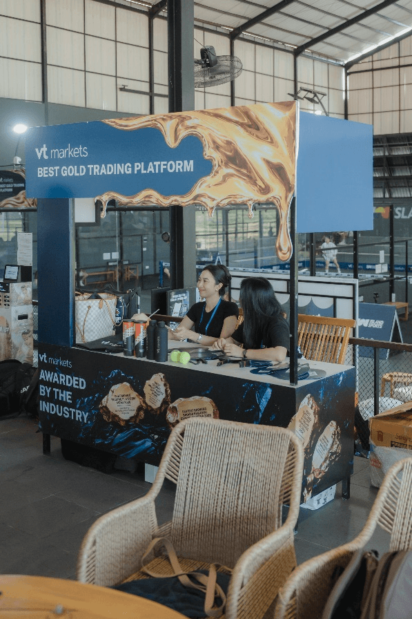 Booth at a trade show two women sit at a VT Markets gold trading platform booth with a blue marble banner reading BEST GOLD TRADING PLATFORM