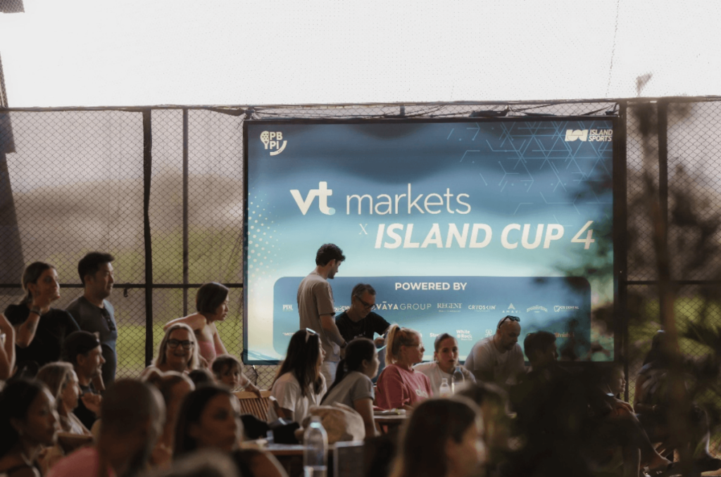 Audience seated at an outdoor event in front of a large VT Markets Island Cup 4 banner on a chain link fence backdrop